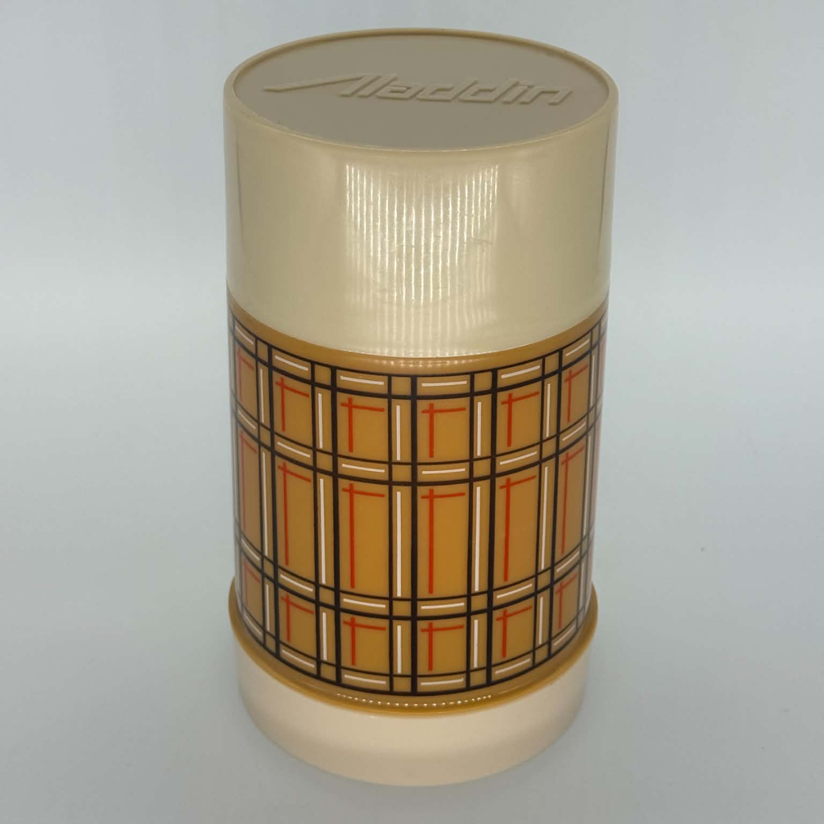 A vintage plastic cylindrical container with a cream-colored ribbed lid and base, featuring a central patterned band with a tan, orange, and black plaid design. The brand name 'Aladdin' is embossed on the lid.
