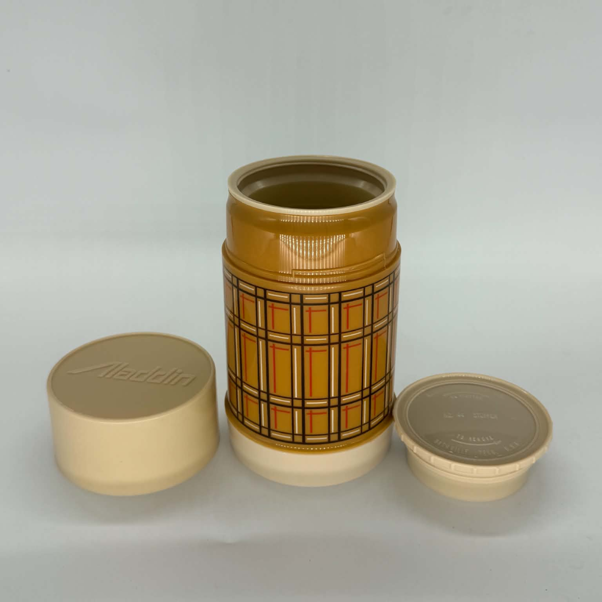 A vintage Aladdin plastic spice jar with a yellow and orange plaid pattern, shown with its cream-colored screw-on lid off, displaying both the inside and the embossed Aladdin logo on the outside.