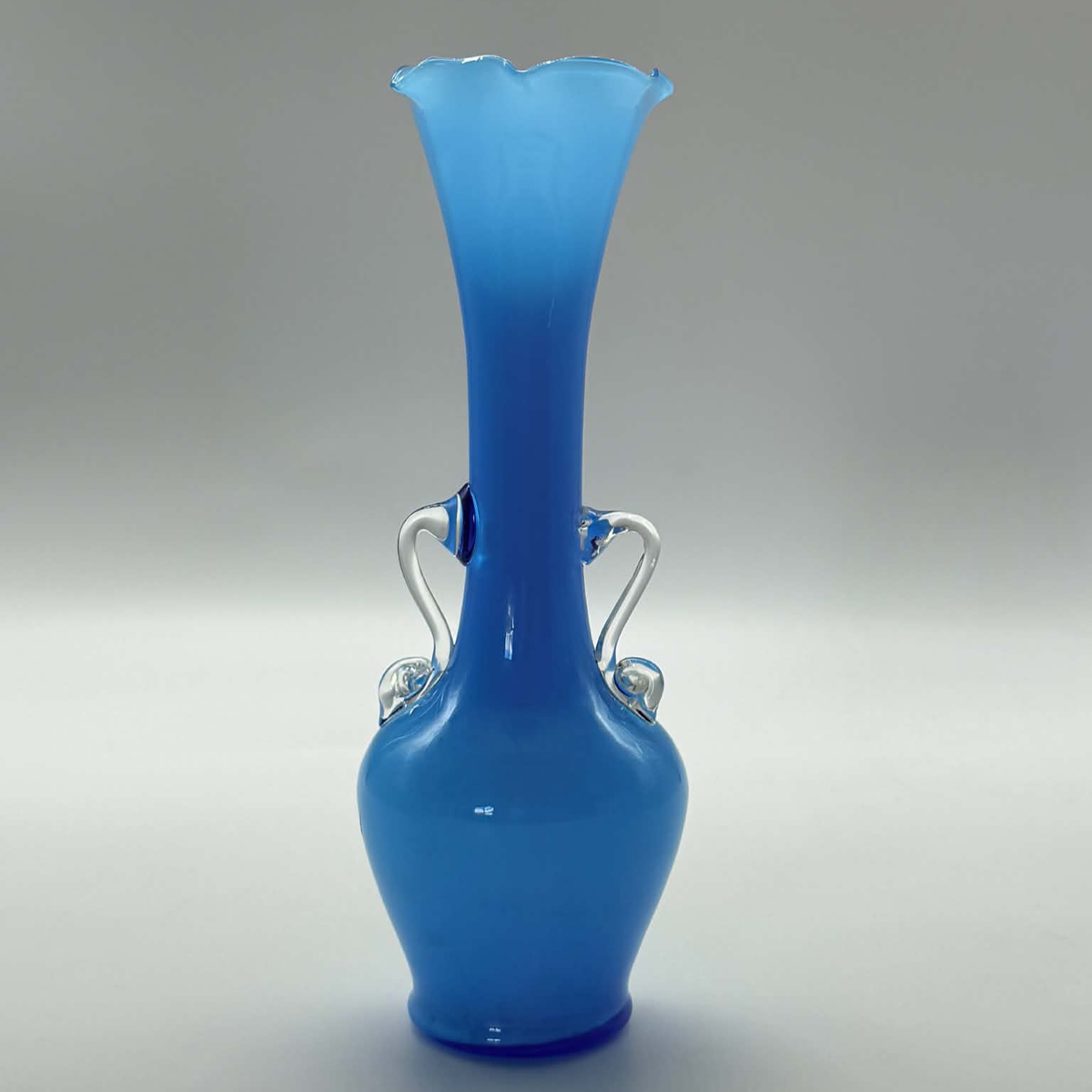 A sky blue glass vase with a flared neck, a rounded base, and two small loop handles, photographed against a neutral gray studio background.