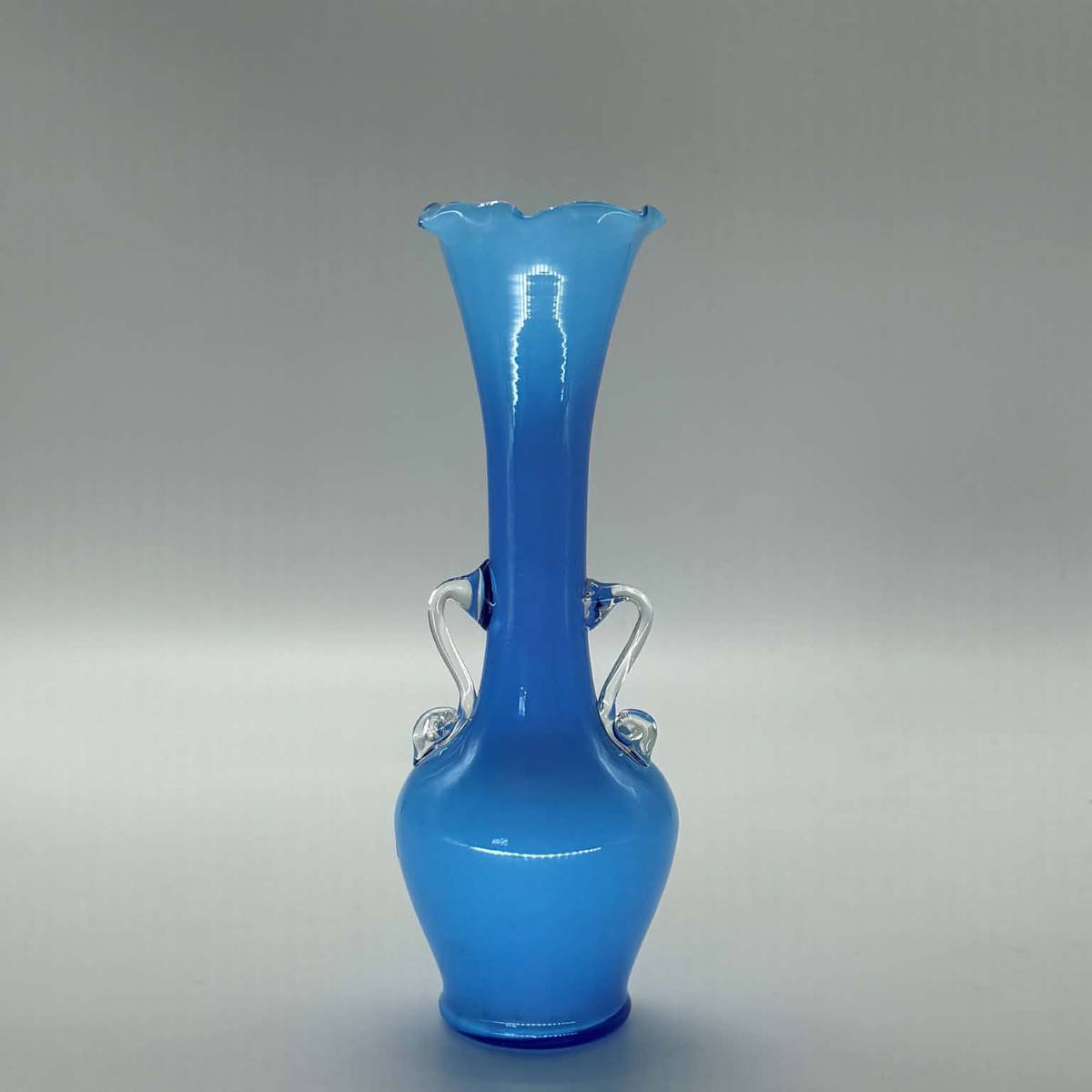 A sky blue glass vase with a flared, scalloped rim, a bulbous base, and two small decorative handles on the sides, photographed against a neutral gray studio background.