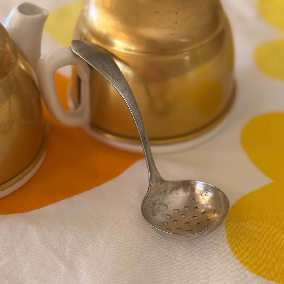 A vintage silver plated tea strainer spoon with a perforated spoon bowl, resting on a white tablecloth with yellow and orange polka dots.
