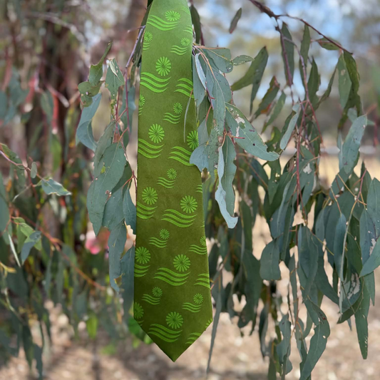 An olive green necktie with a repeating pattern of green sunflowers and wavy lines, hanging from a tree branch surrounded by eucalyptus leaves.