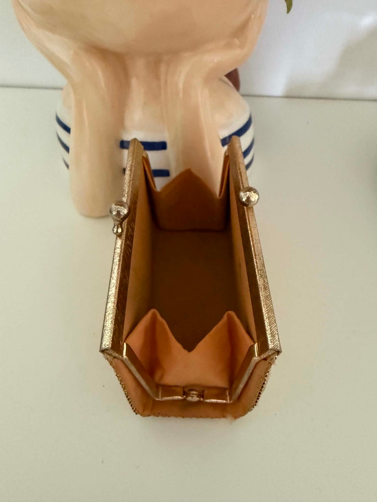A small rectangular clutch purse covered in gold toned metallic mesh like, with a gold-tone frame and clasp, resting on a white surface next to decorative ceramic objects.