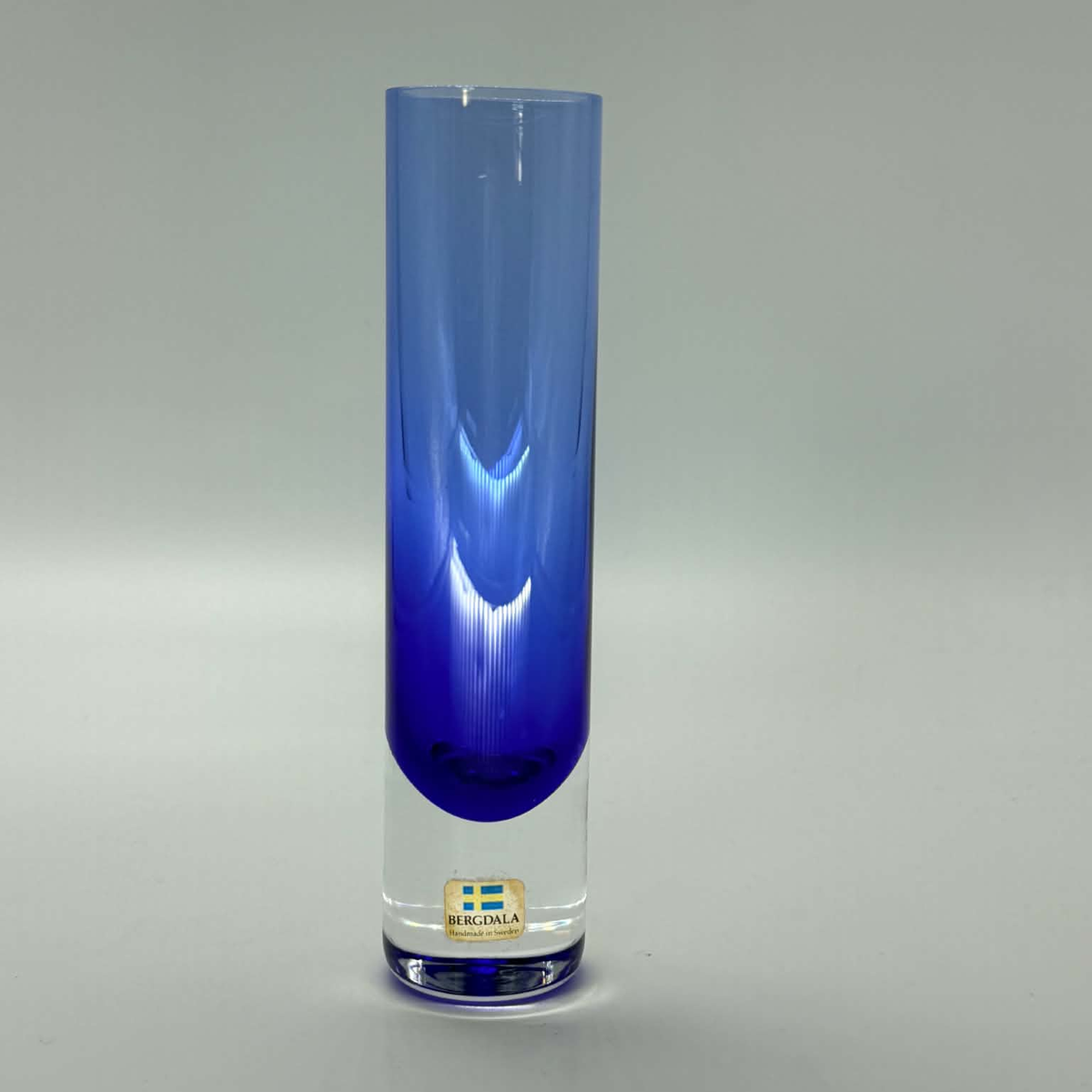 A tall, cylindrical glass vase with a gradient color transition from clear glass at the top to a deep cobalt blue at the bottom. A small gold-colored Bergdala made in sweden brand label is visible near the base.