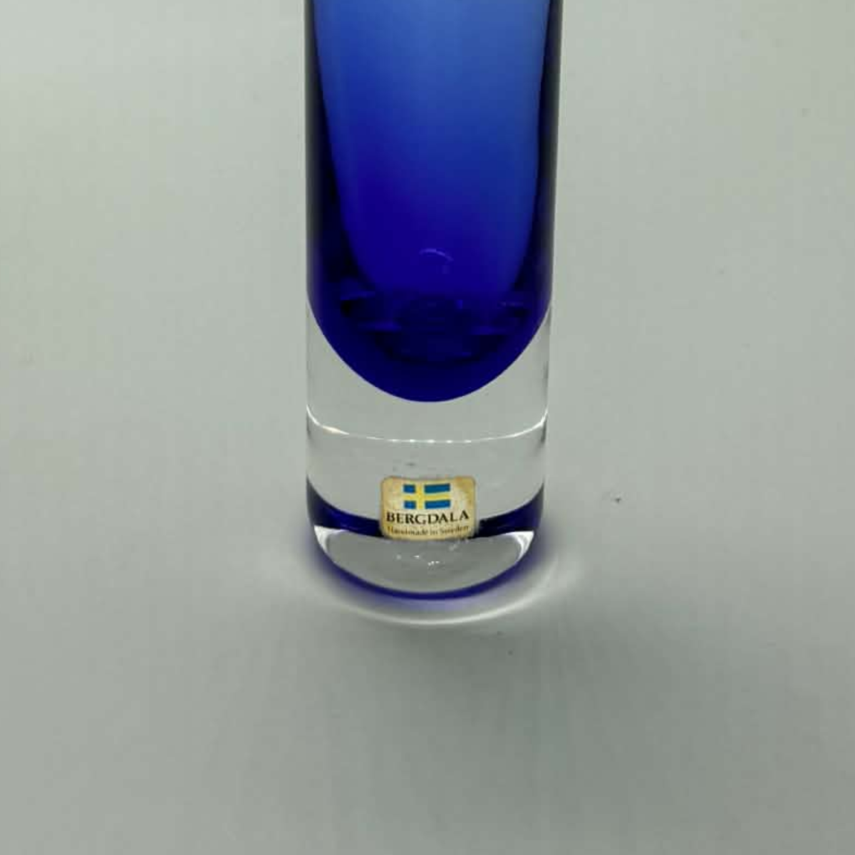 A tall, cylindrical glass vase with a gradient color transition from clear glass at the top to a deep cobalt blue at the bottom. A small gold-colored Bergdala made in sweden brand label is visible near the base.