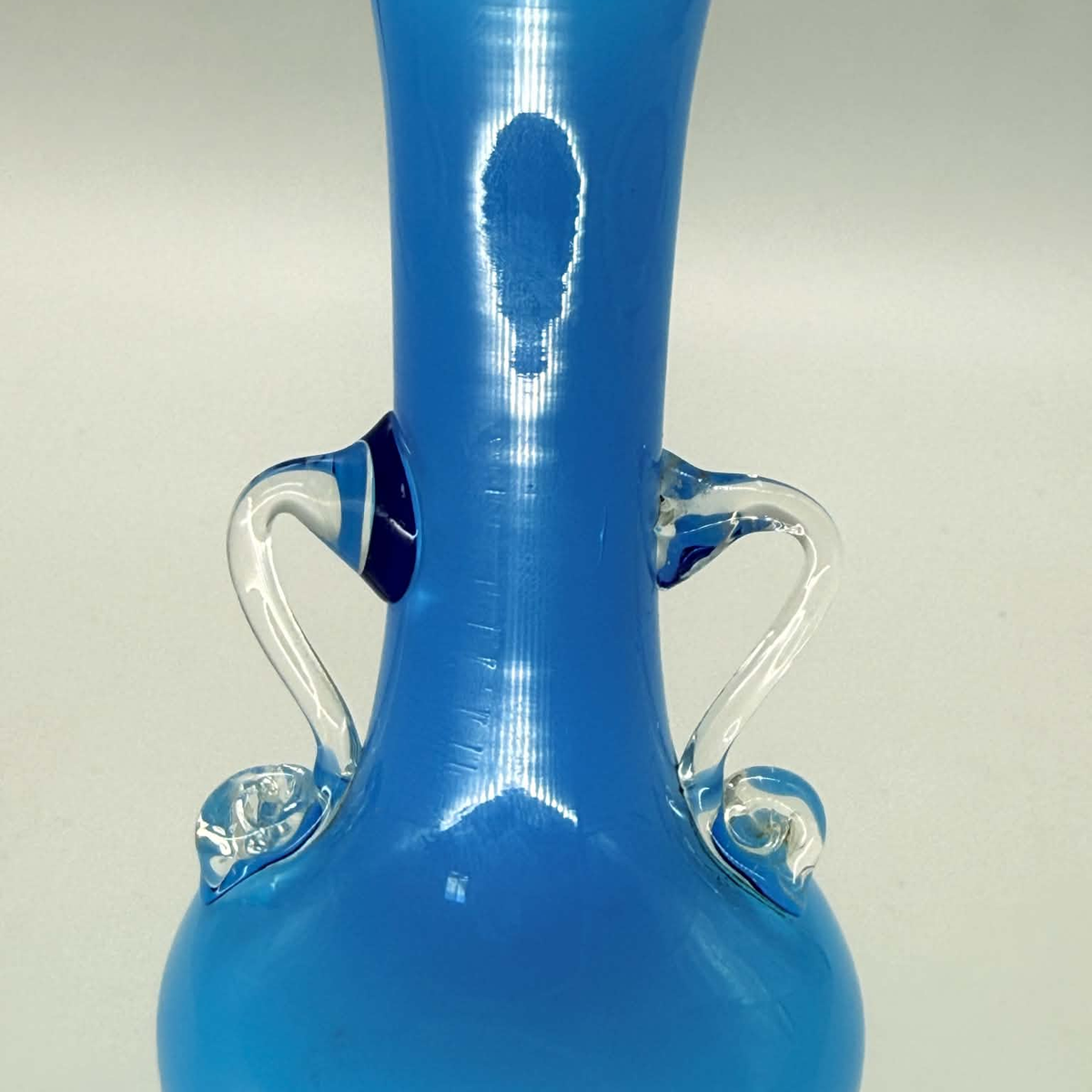 A blue glass vase with a flared round base, a narrow neck, and two clear glass handles, shown against a light neutral background.