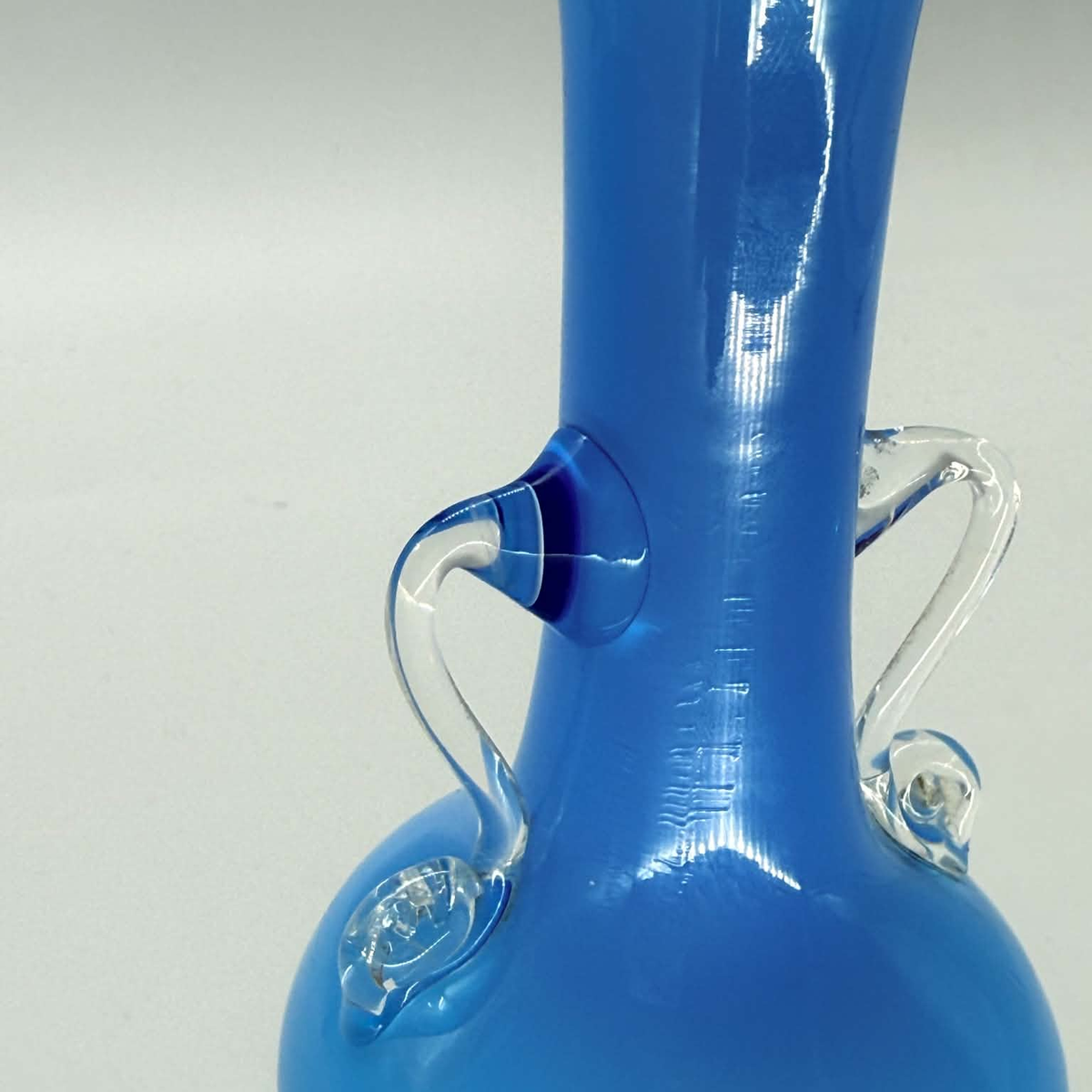 A close-up of a sky blue glass vase with clear glass handles and a flared base, photographed against a light gray background.