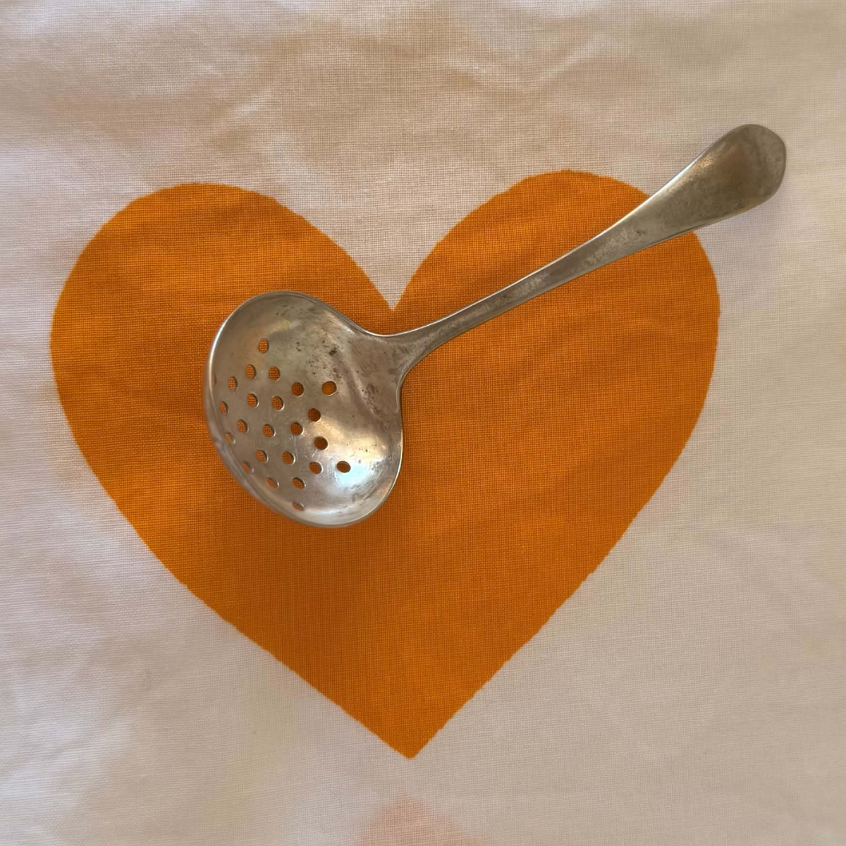 A vintage silver plated metal ladle with perforated holes rests on a white fabric napkin printed with a large orange heart.