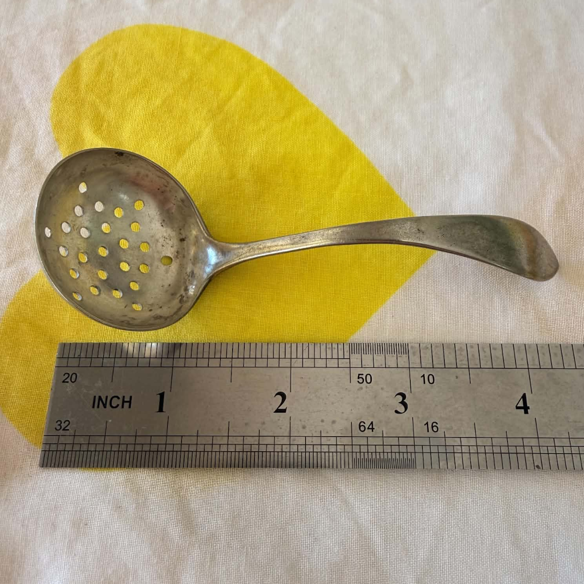A vintage metal strainer spoon with a perforated bowl, shown next to a metal ruler for scale on a white fabric background with a yellow heart-shaped cutout.