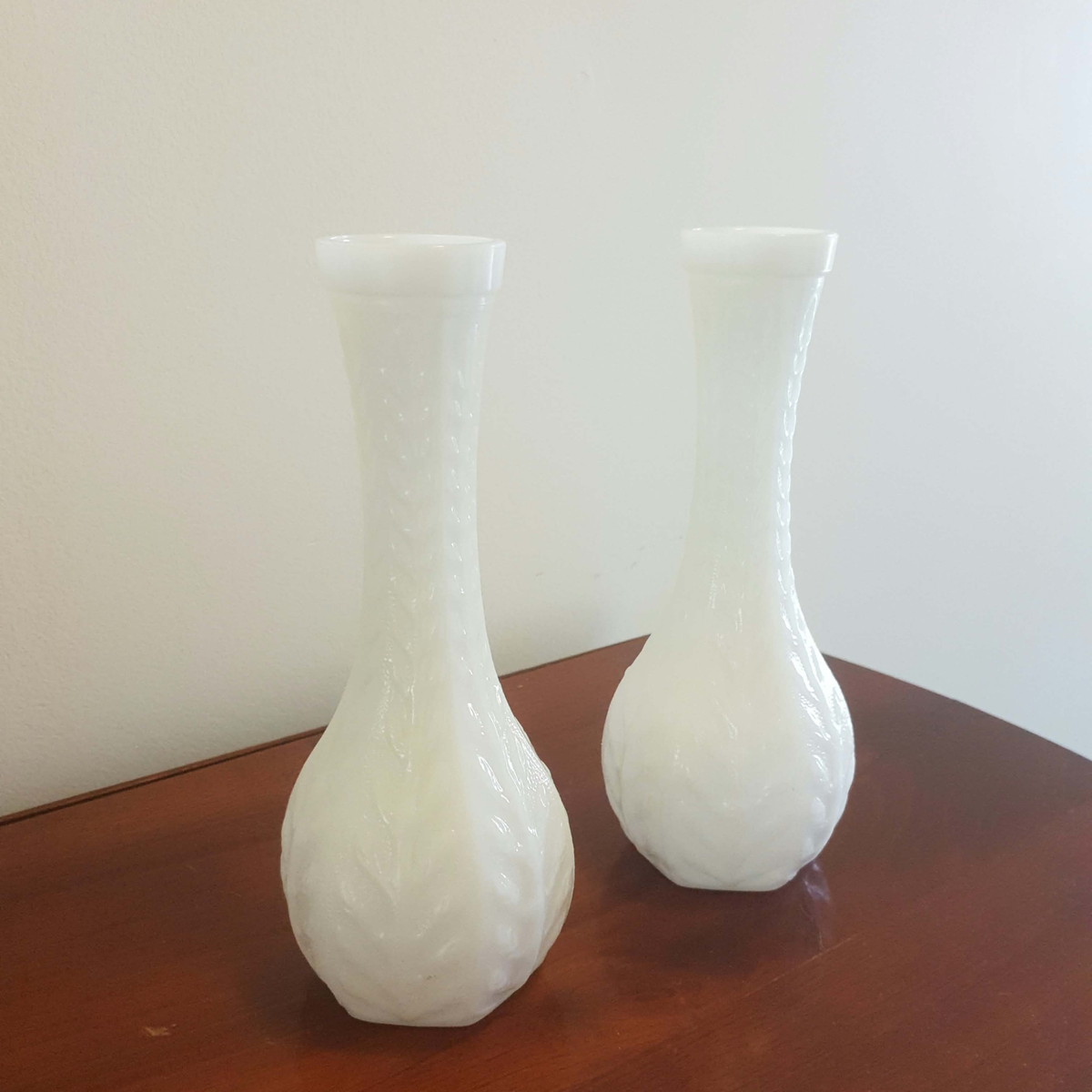 Two white milk glass vases with textured, embossed floral patterns, displayed on a dark wood table against a plain off-white wall.