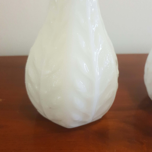 A close-up of a glossy white teardrop-shaped vase or decorative object with a leaf-like embossed texture, resting on a wooden surface against a light wall.
