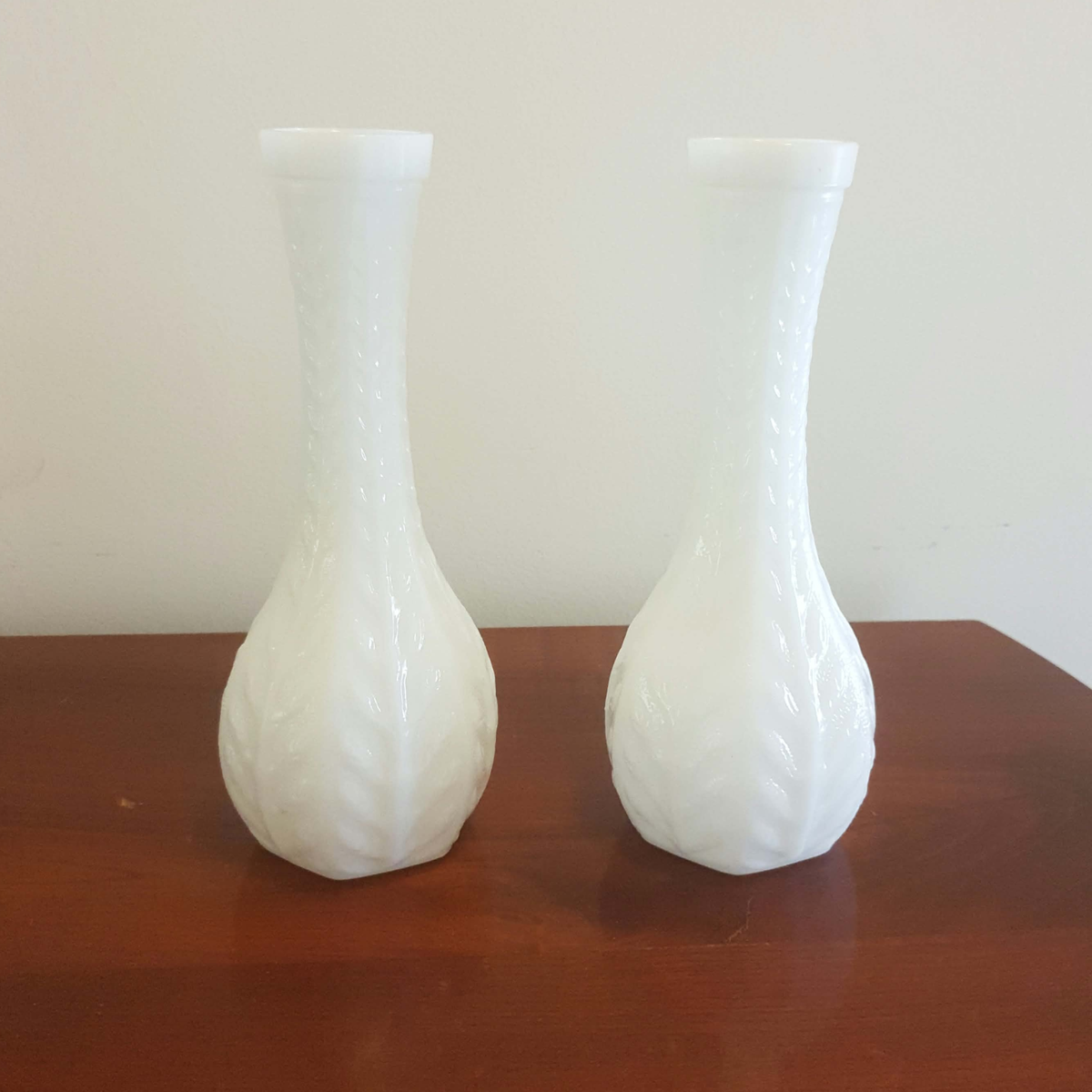 Two white milk glass vases with a textured, embossed leaf pattern, displayed side-by-side on a wooden table against a plain light wall.