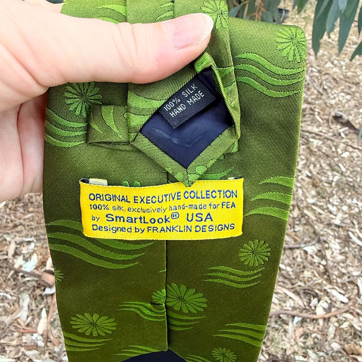 A close-up of a folded olive green silk tie with a green floral and wave pattern, held by a hand. The tie features a yellow paper label reading 'ORIGINAL EXECUTIVE COLLECTION 100% SILK (originally a handmade for FEA by SmartLook® USA Designed by FRANKLIN DESIGNS'.