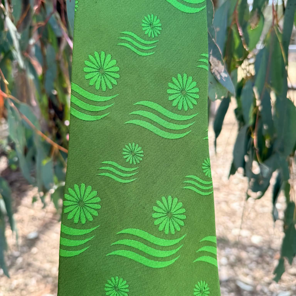 A green necktie with a matching green floral and wave pattern, photographed outdoors against a blurred background of eucalyptus leaves and sunlight.