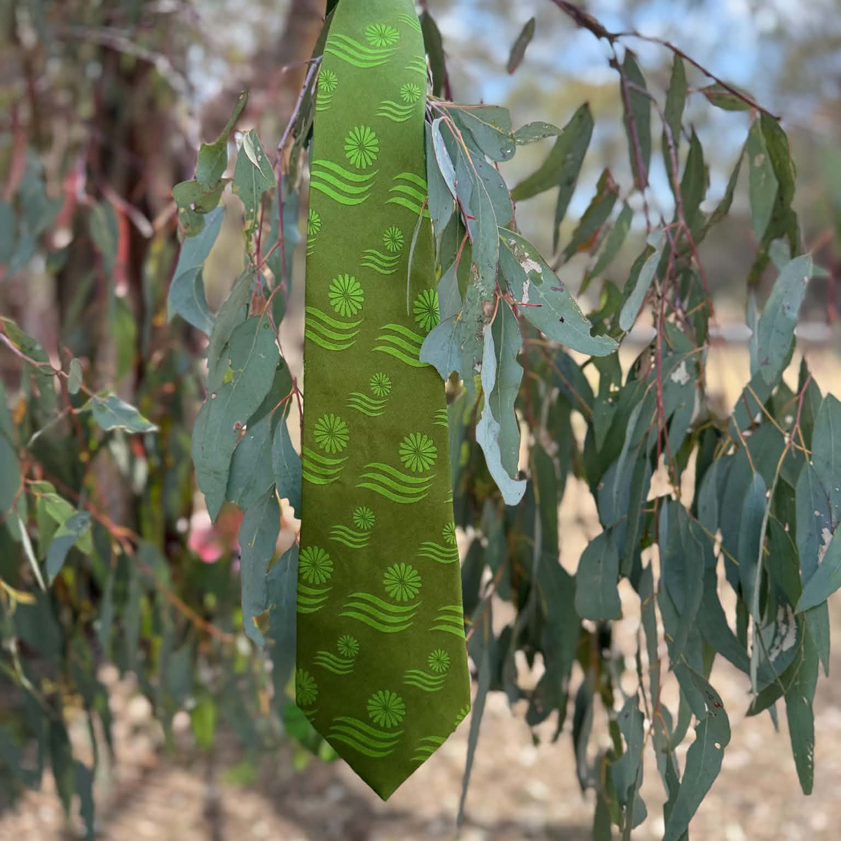 An olive green necktie with a repeating pattern of green sunflowers and wavy lines, hanging from a tree branch surrounded by eucalyptus leaves.