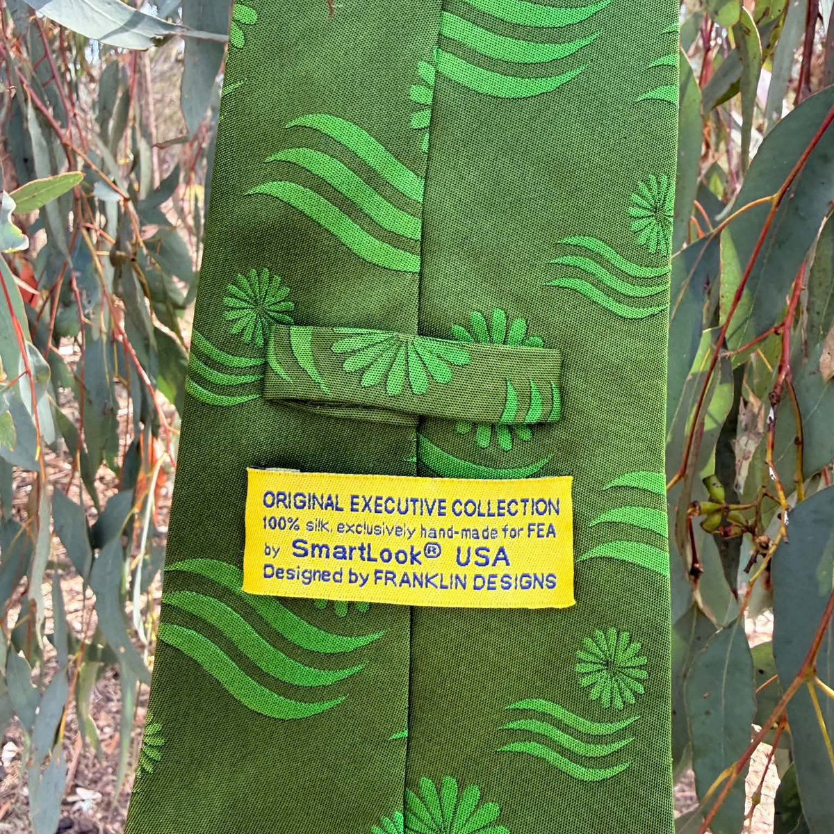 A green silk necktie with a lighter green abstract wavy and floral pattern, featuring a yellow paper label that reads 'ORIGINAL EXECUTIVE COLLECTION 100% silk: exclusively hand-made for FEA by Smart.look® USA Designed by FRANKLIN DESIGNS'.