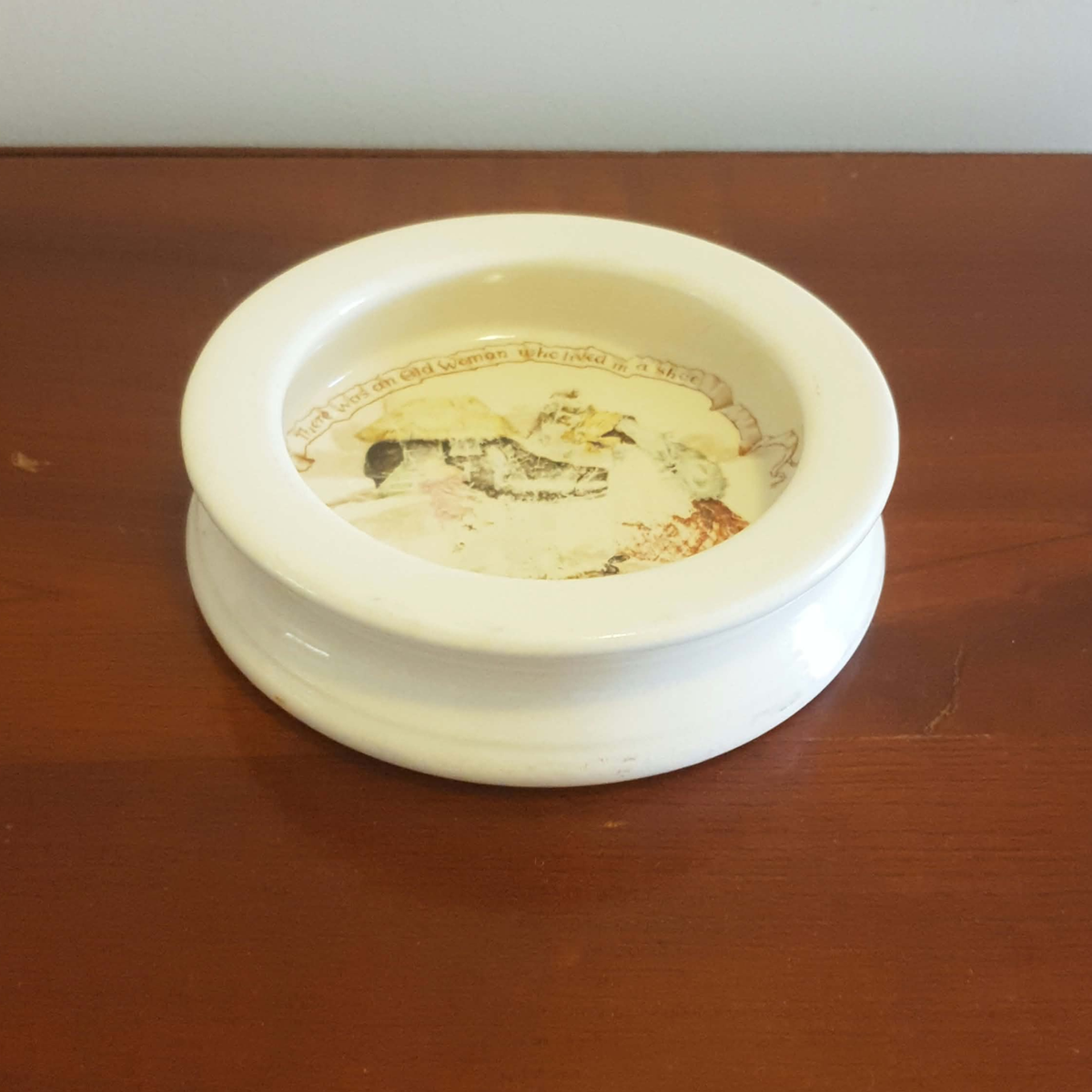 A white ceramic ashtray with a tiered base and a printed religious illustration inside, sitting on a dark brown wooden table.