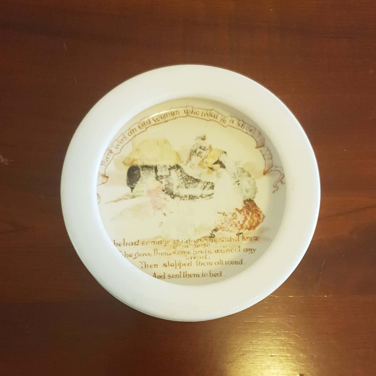 A round white ceramic dish or shallow bowl with a printed illustration and text, photographed from above on a dark wooden surface.
