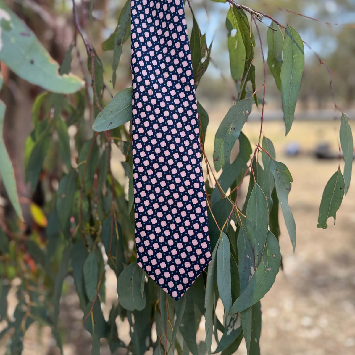 A patterned necktie hanging from a leafy branch outdoors, featuring a navy blue base with a repeating small pink and white floral motif.