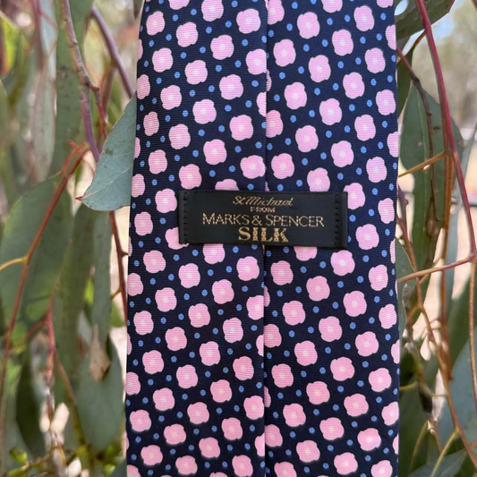 A silk necktie with a navy blue base, pink floral motifs, and light blue polka dots, hanging outdoors among green leaves. The brand label reads 'Sutton & Spence' and 'Marks & Spencer Silk'.