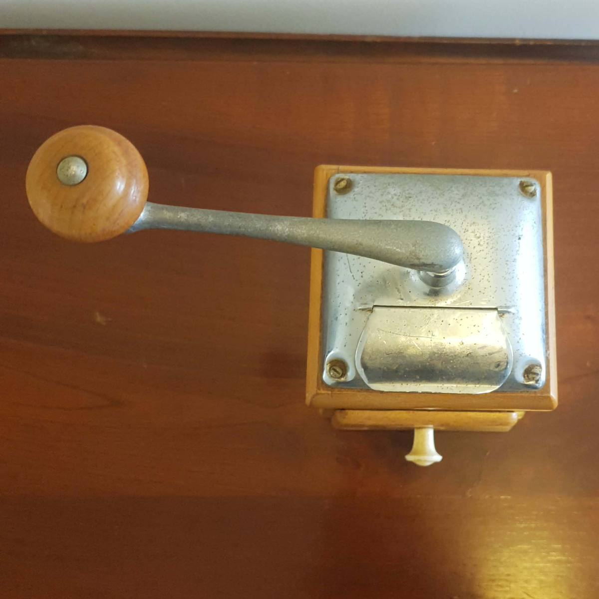 A vintage manual coffee grinder with a wooden handle and a square metal body, resting on a polished wooden surface.