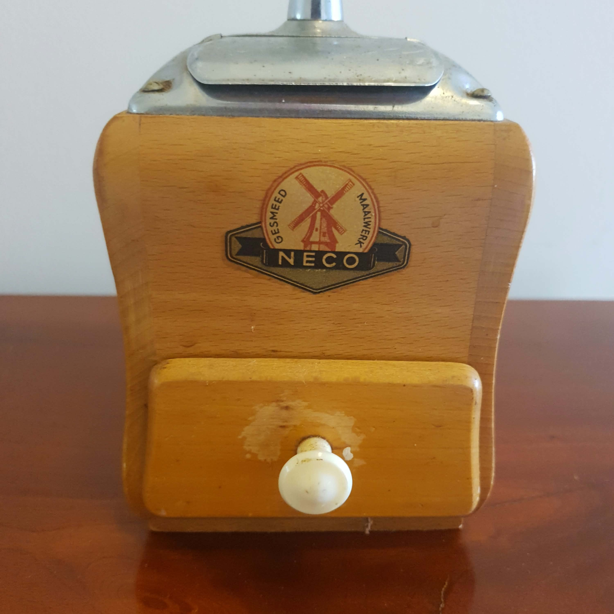 A vintage wooden NECO grain mill with a metal top and a white plastic or enamel button on the front, resting on a wooden table against a plain light background.
