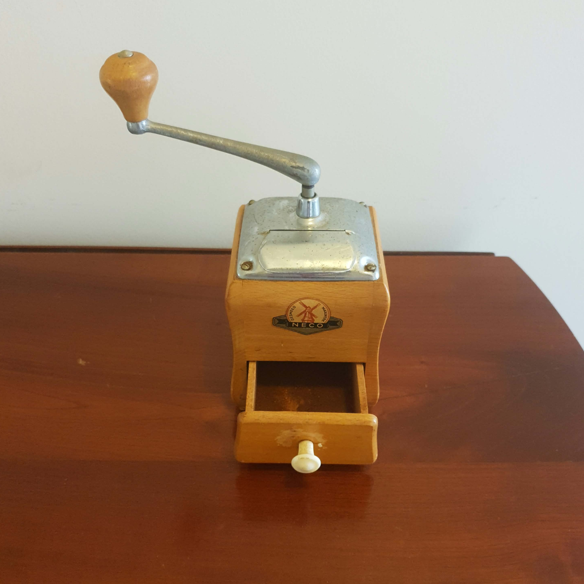 A vintage wooden hand-crank coffee grinder with a metal lever and a small drawer, set on a wooden table against a plain wall.