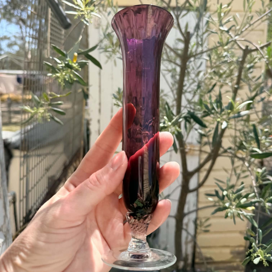 A hand holding a tall, flared purple glass vase with a textured base, set against a blurred outdoor background of green plants and a fence.