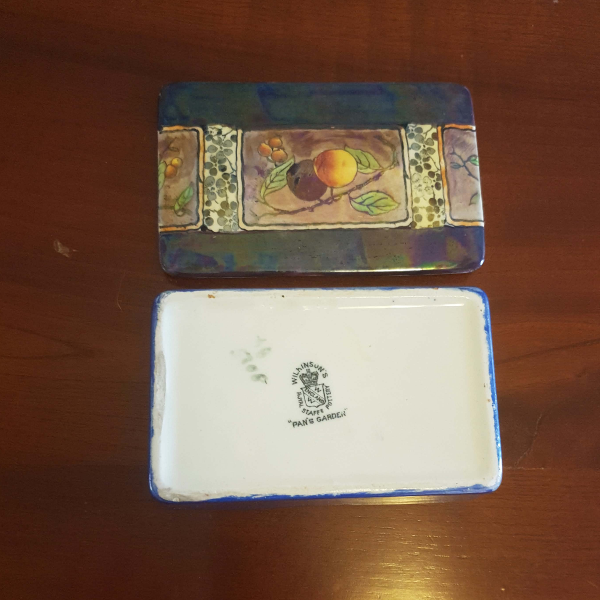 A rectangular ceramic dish or box shown from two angles on a wooden table. The top view shows a decorative multicolor floral and fruit pattern, while the bottom view shows a white interior with a blue rim and a printed crest reading 'Peach Gardens'.