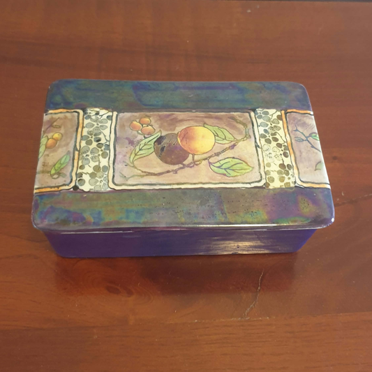 A rectangular decorative ceramic box with a painted fruit motif on the lid, resting on a wooden surface. lustre finish