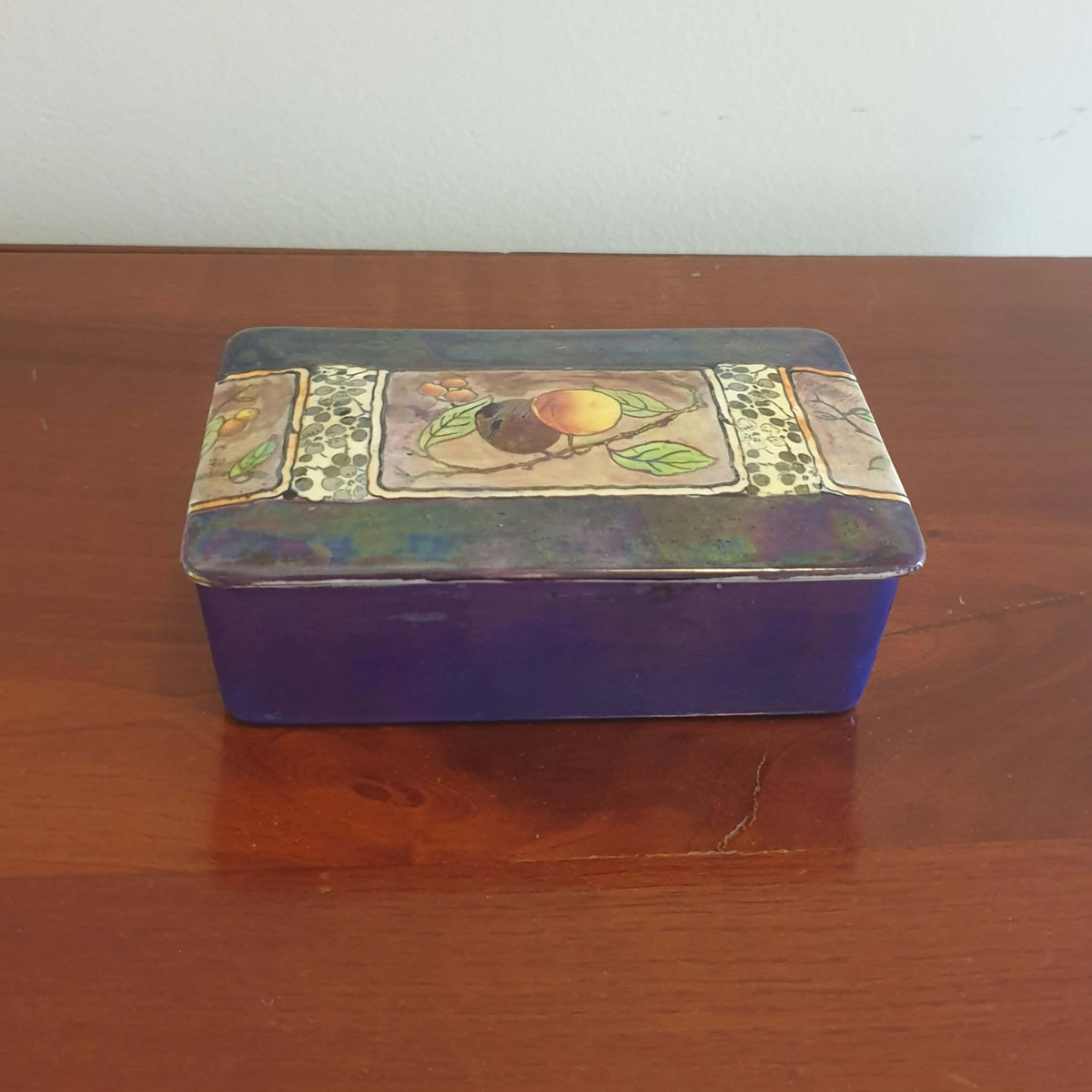A vintage rectangular ceramic box with a purple base and a decorative lid featuring a fruit illustration and floral borders, resting on a wooden table against a plain wall.
