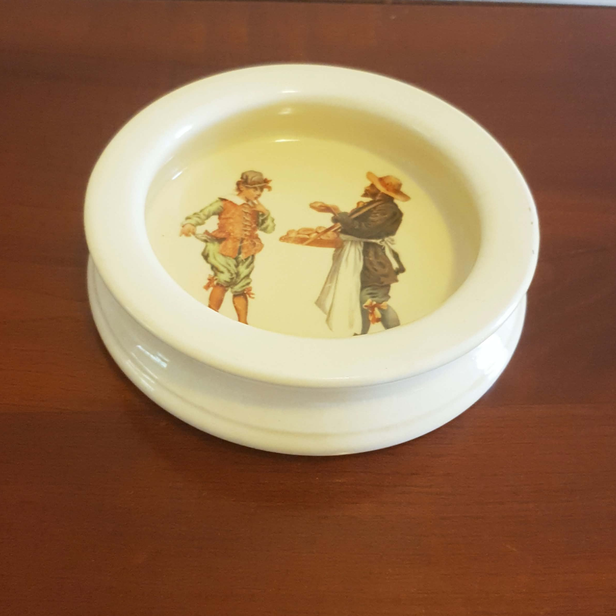 A round white ceramic dish with a raised rim and a printed illustration of two historical figures on the inside, set on a dark brown wooden surface.
