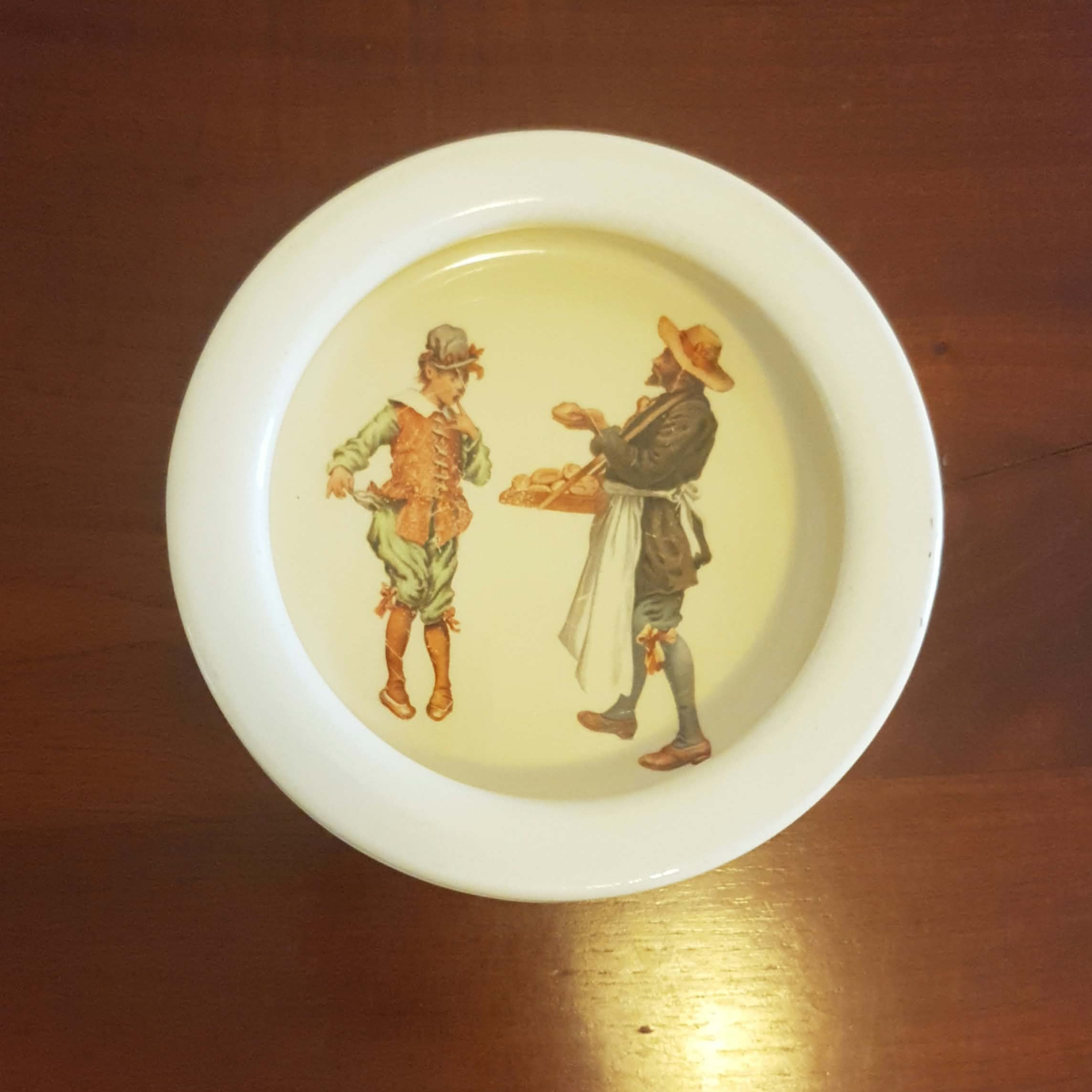 A white ceramic bowl with a vintage illustration of two men in historical clothing on a light cream background, shown on a dark wooden surface.