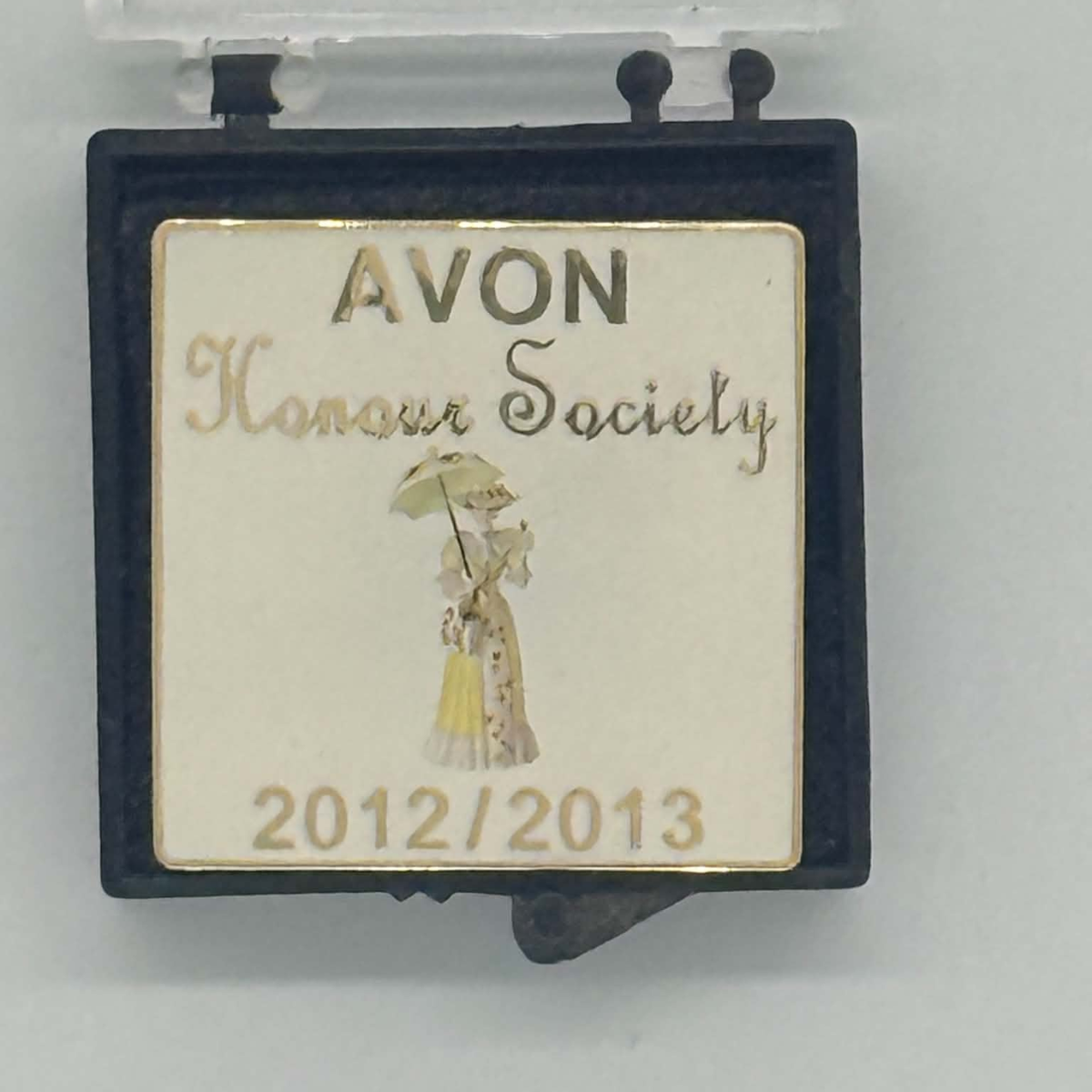 A square black-framed Avon Honour Society pin or badge with an off-white center panel, gold-toned lettering, and a small illustration of a woman holding an umbrella. The text reads 'AVON Honour Society' and '2012/2013'.