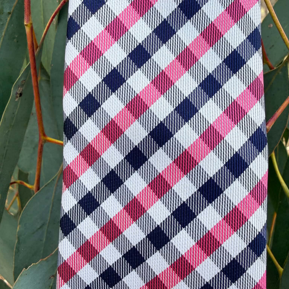 A close-up of a necktie featuring a diagonal gingham check pattern in pink, navy blue, and white, photographed outdoors against green leaves.
