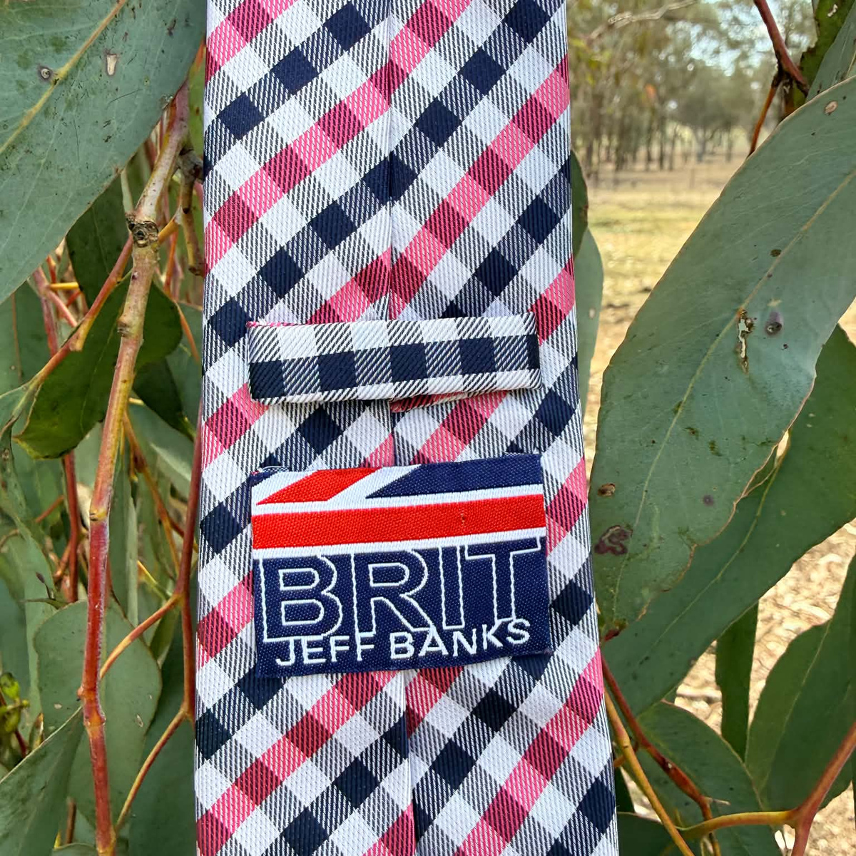A necktie with a pink, white, and navy blue gingham pattern, hanging outdoors among green leaves. The tie features a matching fabric loop and a rectangular patch with the text 'BRIT JEFF BANKS' and a Union Jack design.
