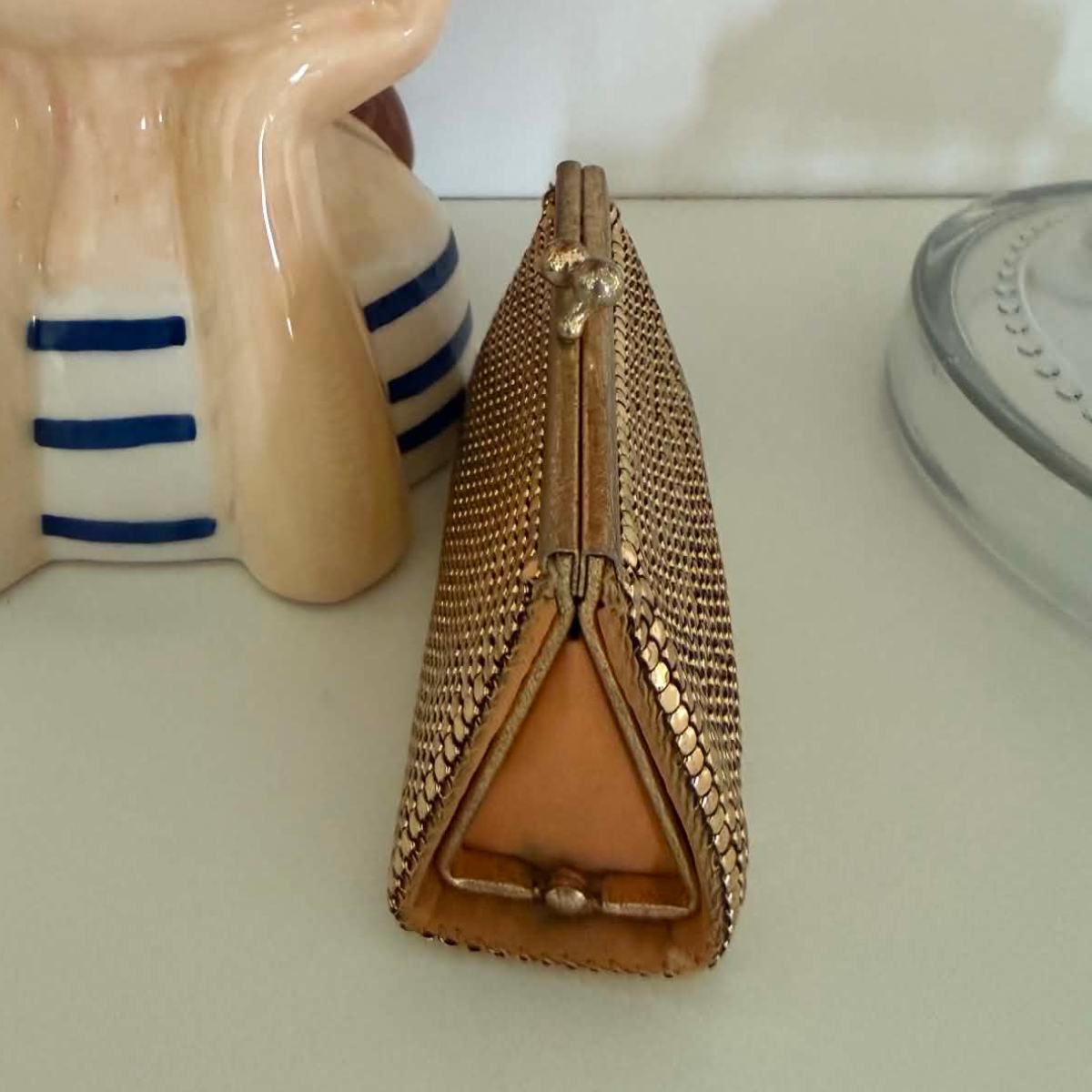 A small rectangular clutch purse covered in gold toned metallic mesh like, with a gold-tone frame and clasp, resting on a white surface next to decorative ceramic objects.