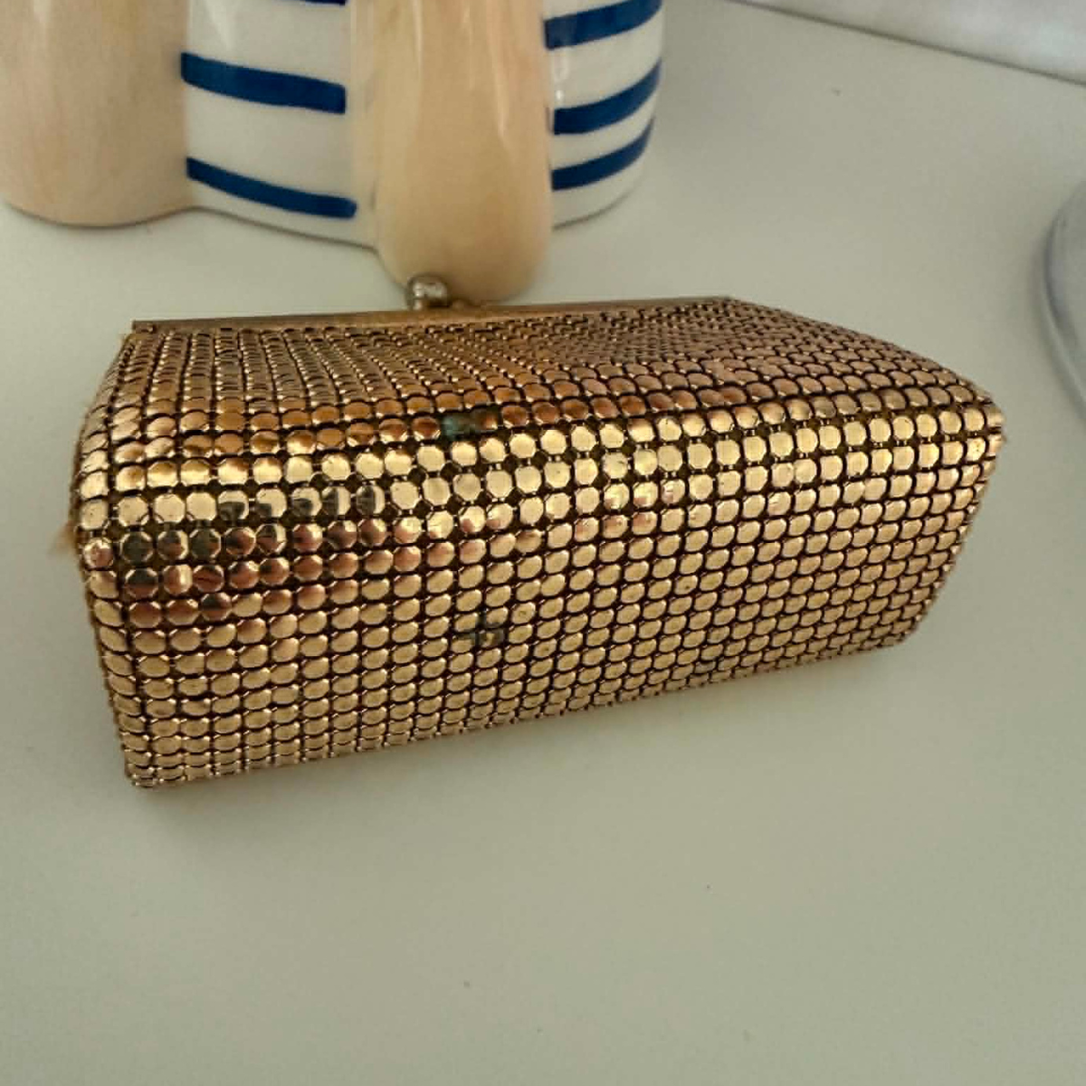 A small rectangular clutch purse covered in gold toned metallic mesh like, with a gold-tone frame and clasp, resting on a white surface next to decorative ceramic objects.