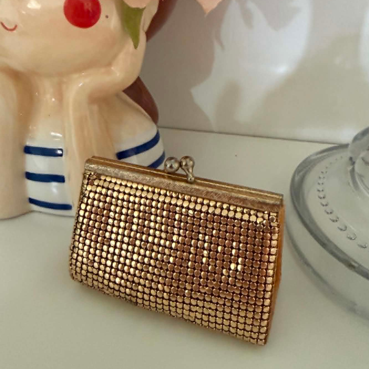 A small rectangular clutch purse covered in gold toned metallic mesh like, with a gold-tone frame and clasp, resting on a white surface next to decorative ceramic objects.