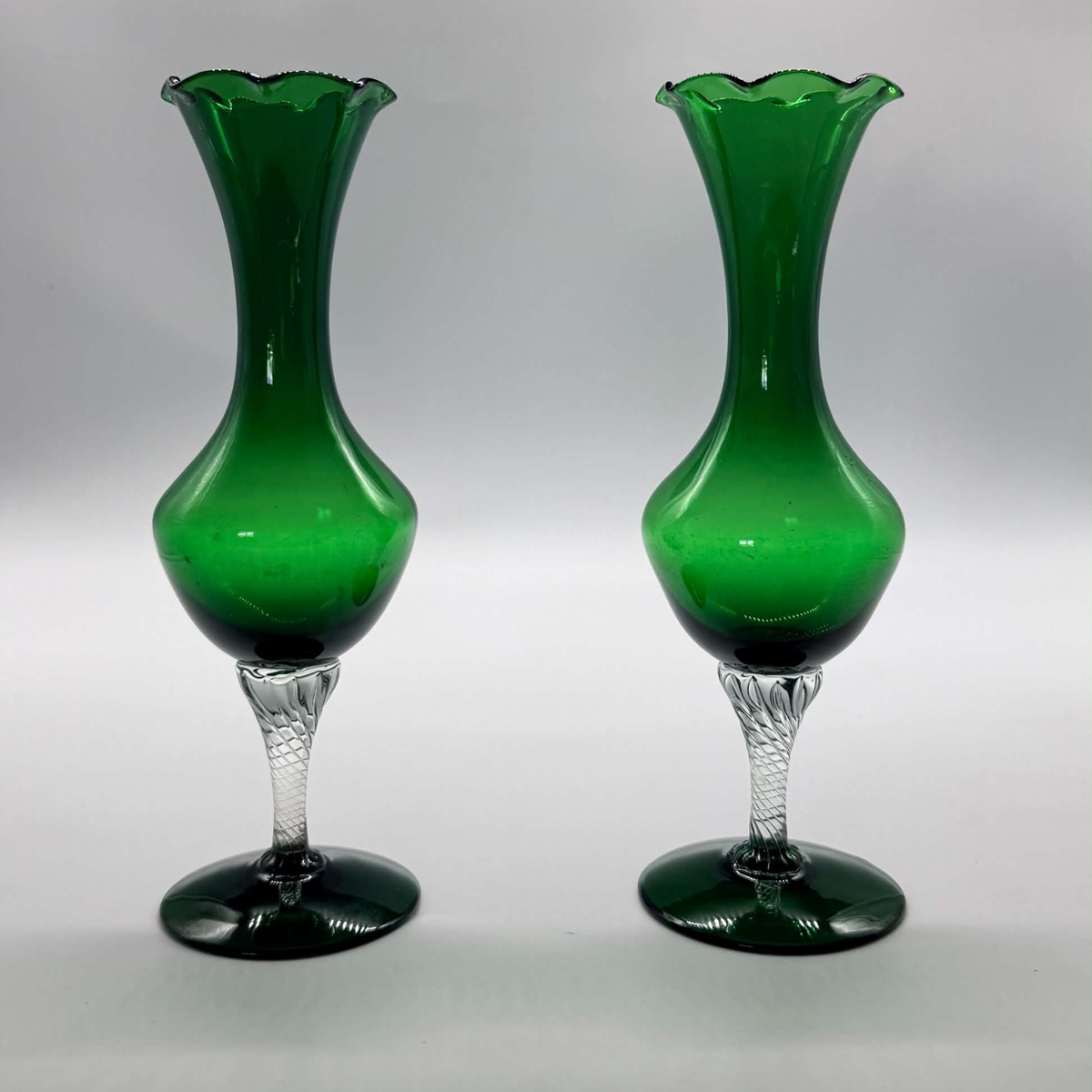 A pair of green glass flower vases with flared scalloped rims, bulbous bodies, and clear faceted stems on round dark green bases, photographed against a neutral gray studio background.