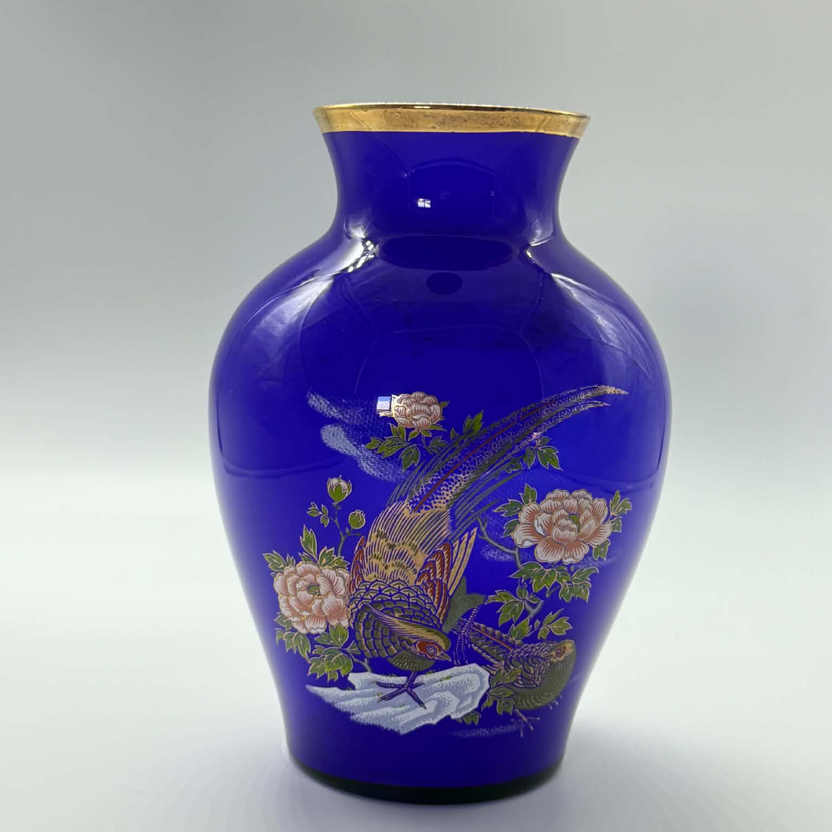A cobalt blue glass vase with a gold rim, decorated with a hand-painted pheasant and pink flowers on a light gray background.