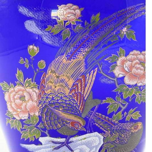 A close-up of a vibrant cobalt blue ceramic vase decorated with hand-painted green leaves, pink flowers, and two detailed pheasant birds.