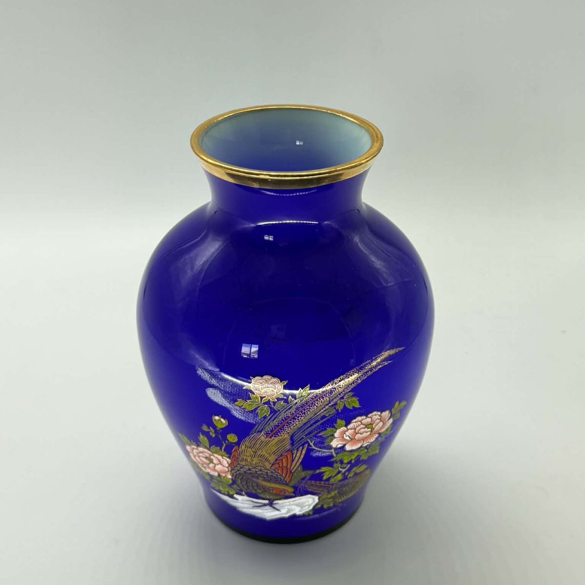 A cobalt blue ceramic vase with a gold rim, decorated with a hand-painted phoenix and floral motif in pink, green, and gold, set against a neutral gray background.
