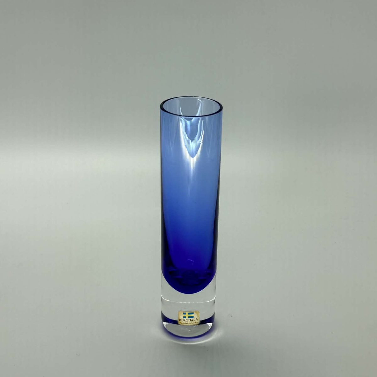 A tall, cylindrical glass vase with a gradient color transition from clear glass at the top to a deep cobalt blue at the bottom. A small gold-colored Bergdala made in sweden brand label is visible near the base.