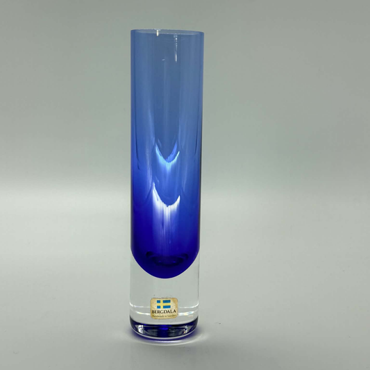 A tall, cylindrical glass vase with a gradient color transition from clear glass at the top to a deep cobalt blue at the bottom. A small gold-colored Bergdala made in sweden brand label is visible near the base.