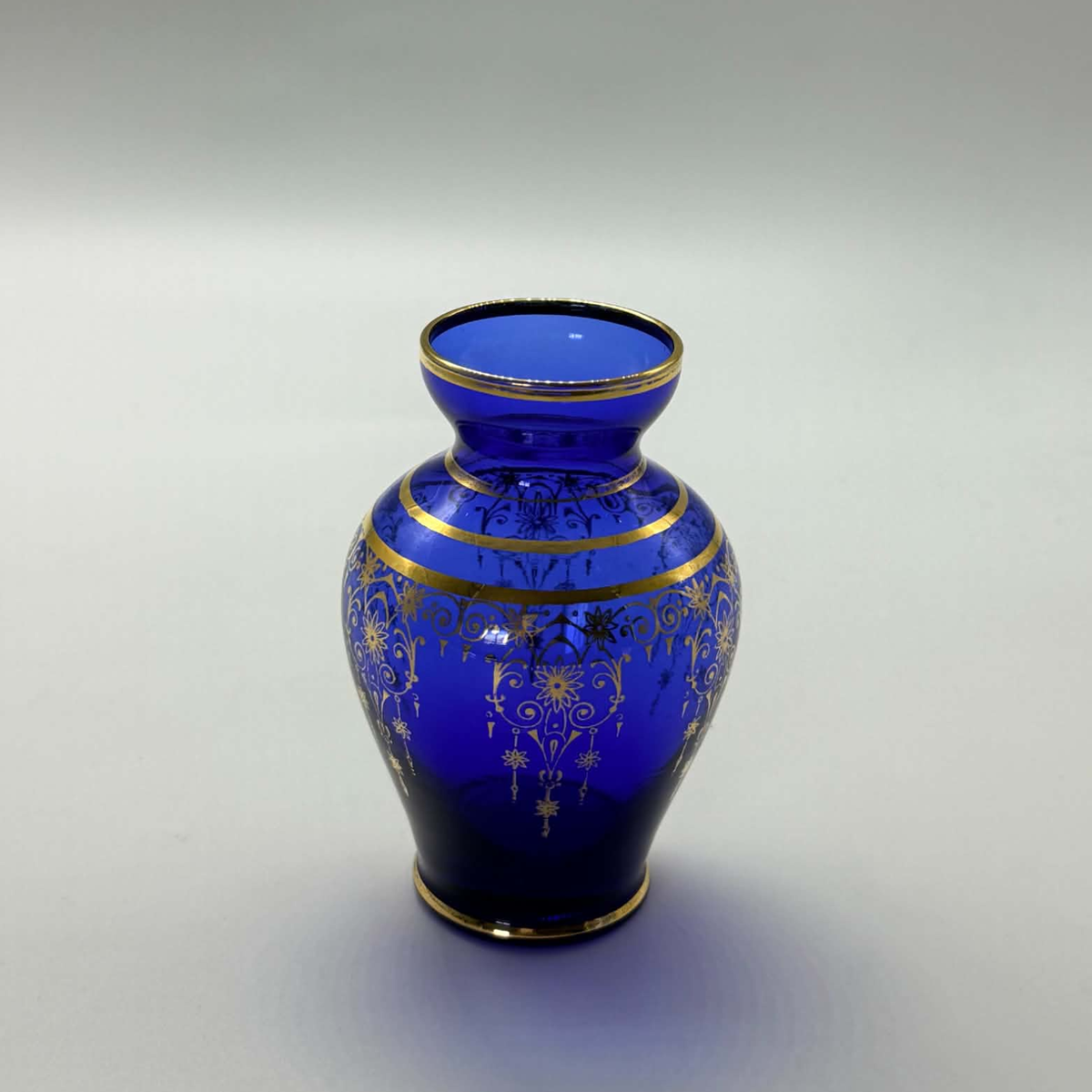 A cobalt blue glass vase with ornate gold floral patterns and gold trim, photographed on a neutral light gray background.