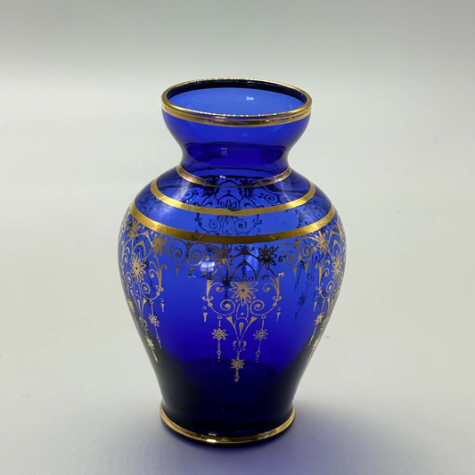 A cobalt blue glass vase with ornate gold filigree patterns and gold trim, photographed on a neutral light gray background.