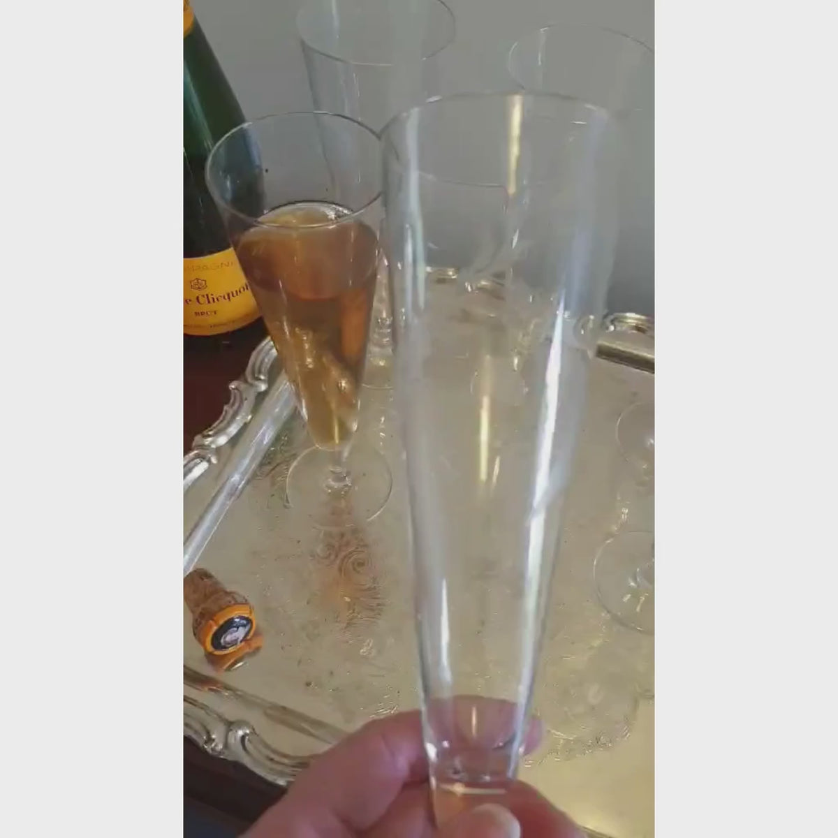 A green Veuve Clicquot champagne bottle with a yellow label stands beside a large ornate silver tray holding six clear champagne flutes, one partially filled with golden champagne, against a plain light wall.