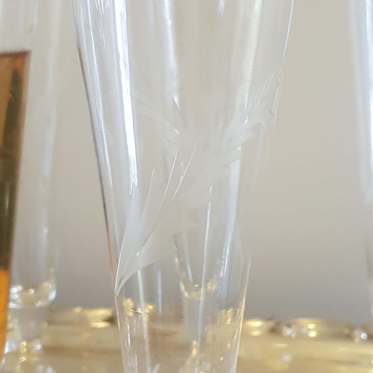 A green Veuve Clicquot champagne bottle with a yellow label stands beside a large ornate silver tray holding six clear champagne flutes, one partially filled with golden champagne, against a plain light wall.