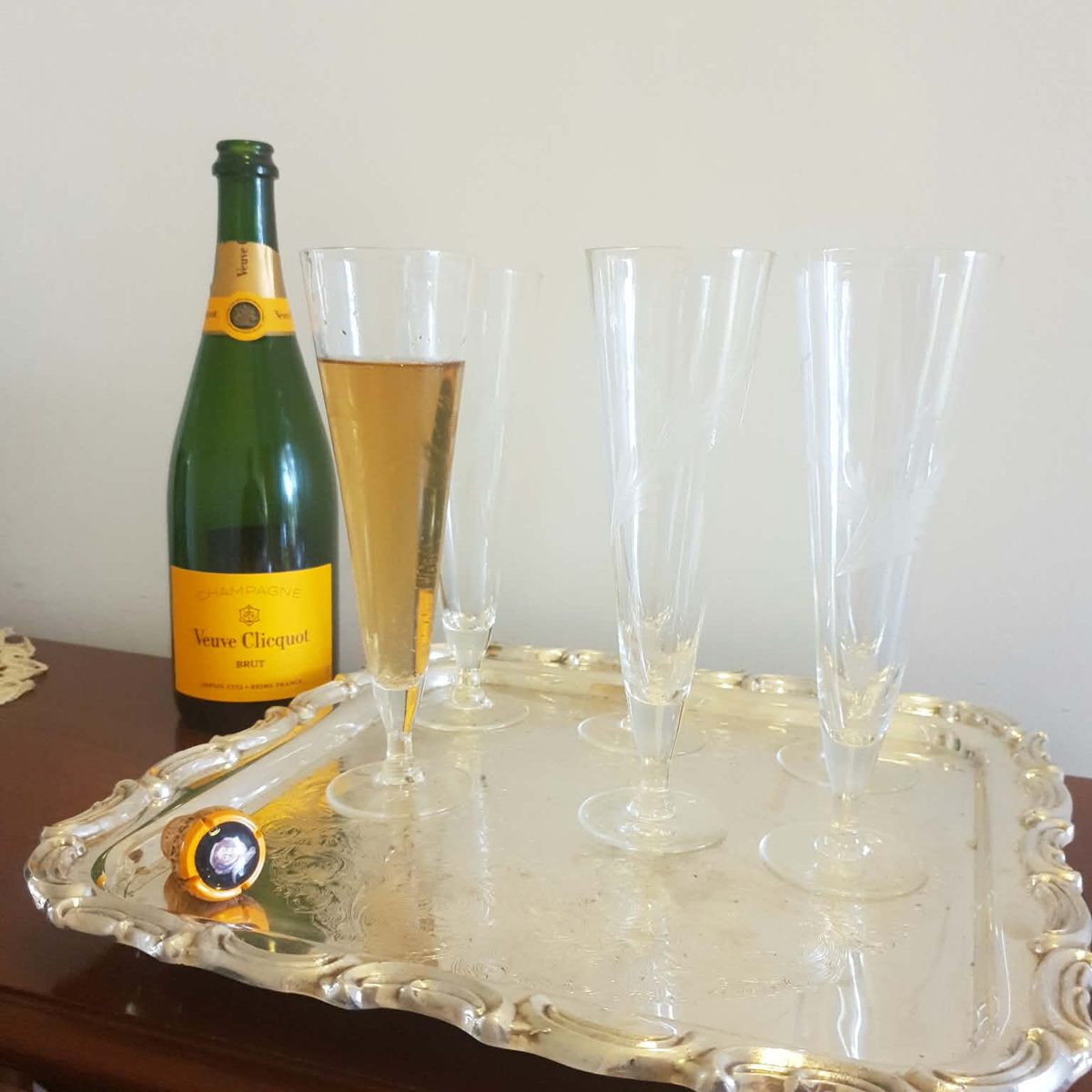 A green Veuve Clicquot champagne bottle with a yellow label stands beside a large ornate silver tray holding six clear champagne flutes, one partially filled with golden champagne, against a plain light wall.